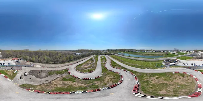 Aerial 360-degree view of Boschertown Go Karts’ winding track in St. Charles, bordered by red and white tires, surrounded by grassy areas, trees, nearby buildings, and a clear blue sky with the sun shining overhead.
