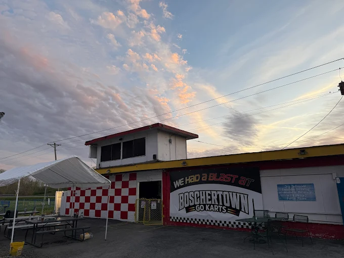 A go-kart building with red and white checkered walls and a “Boschertown Go Karts” sign stands under a wide St. Charles sunset sky, with picnic tables and a canopy in the foreground.