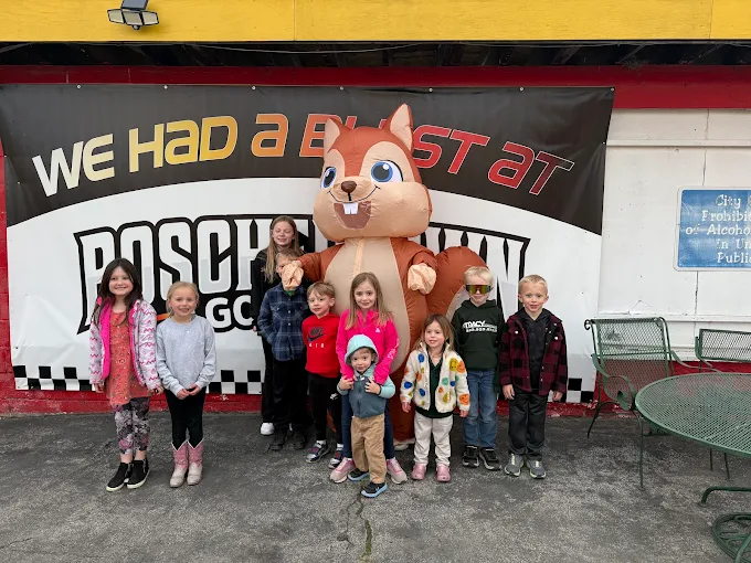 A group of children stands smiling with a person in a large chipmunk costume in front of a banner reading We Had a Blast at POSCH, near the outdoor chairs and tables at Boschertown Go Karts in St. Charles.