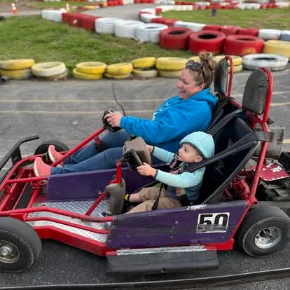 A smiling woman and a small child ride together in a red and purple go-kart at Boschertown Go Karts in St. Charles, both holding the steering wheel on an outdoor track surrounded by colorful tires.