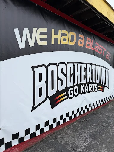 A large outdoor banner reads “WE HAD a BLAST at BOSCHERTOWN GO KARTS,” featuring a black, white, and red design with a checkered border. The banner promotes Go Karts fun in St. Charles and is attached to a wall near a paved surface.