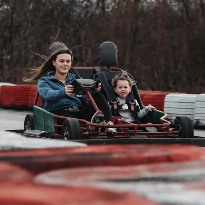 Two young girls ride together in a go-kart at Boschertown Go Karts in St. Charles, their hair blowing in the wind as they race past red and white barriers lining the outdoor track.