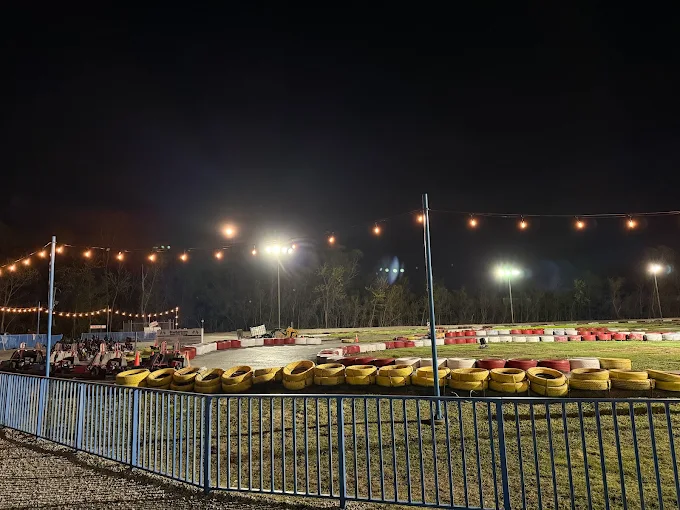 At Boschertown Go Karts in St. Charles, a go-kart track comes alive at night under string lights and floodlights, with yellow tires marking the curves. A blue fence lines the front, while trees stand quietly in the background.