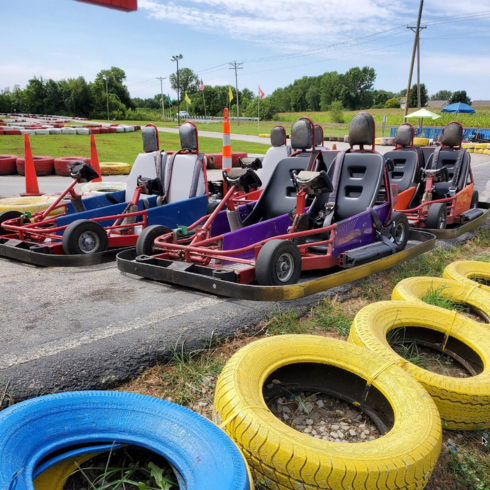 Colorful go-karts are parked side by side on an outdoor track at Boschertown Go Karts in St. Charles, with yellow and blue tires as barriers, surrounded by grass, trees, and distant fencing under a partly cloudy sky.