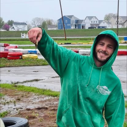 A smiling man in a green hoodie holds up a small fish with two fingers at Boschertown Go Karts on a cloudy day in St. Charles. Colorful tires and houses add charm to the go-kart track in the background.