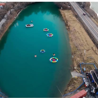 Aerial view of people relaxing on colorful floating rings and a boat in a turquoise lake beside a road near St. Charles, with trees and grassy banks surrounding the water—just a short drive from Boschertown Go Karts fun.