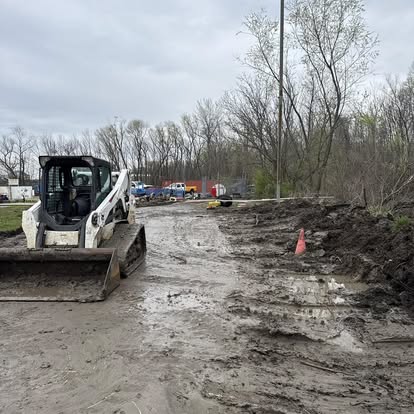 A white skid steer loader sits on a muddy construction site near a dirt road in St. Charles, with trees and parked vehicles in the background. A single orange traffic cone is on the muddy ground to the right.