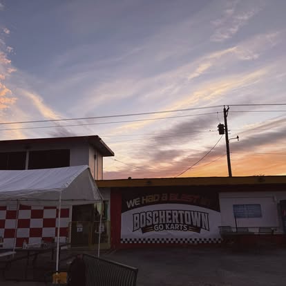 Outdoor view of Boschertown Go Karts in St. Charles at sunset, with a sign reading “We had a blast!”, a white canopy tent, red and white checkered wall, and colorful sky with wispy clouds above.