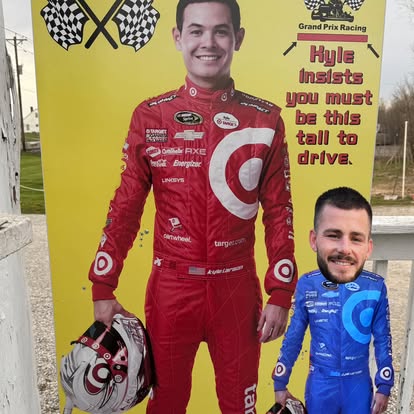 A person stands next to a life-size racing driver cutout at Boschertown Go Karts in St. Charles, their head poking through the face hole for a funny photo. The sign reads, Kyle insists you must be this tall to drive, with checkered flags above.