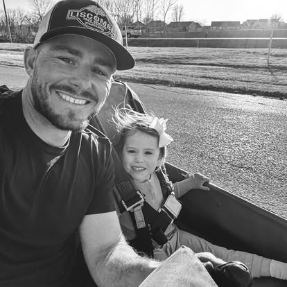 A man and a young girl, both smiling, take a selfie while riding in an open Go Kart on a sunny day at Boschertown Go Karts in St. Charles. The girl wears a large bow and is strapped in. Houses and trees are visible in the background.