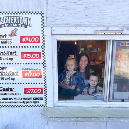 A woman and two young children smile at the Boschertown Go Karts ticket window in St. Charles. Next to them, a price board lists go-kart ride options, set against a white wooden wall in the background.