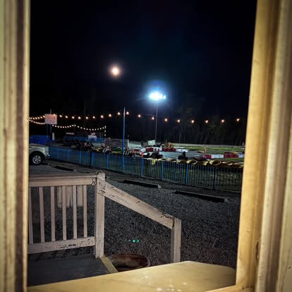 A nighttime view through a window shows the Boschertown Go Karts racetrack in St. Charles, aglow with string lights, parked cars, and a wooden railing in the foreground under a moonlit sky.