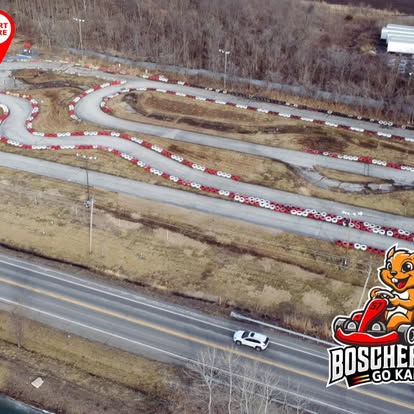 Aerial view of a winding outdoor go-kart track in St. Charles, surrounded by bare trees and a road with a white car. The Boschertown Go Karts logo with a cartoon mascot appears in the lower right corner.