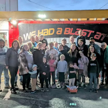 A large group of people, including children and adults, pose and smile together for a photo in front of a sign that reads We Had a Blast At Boschertown Go Karts. The setting appears to be outdoors in St. Charles in the evening.