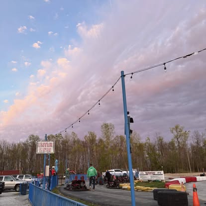 At Boschertown Go Karts in St. Charles, people gather near go karts on the track at sunset, with string lights overhead and trees in the background under a colorful sky streaked with pink clouds.