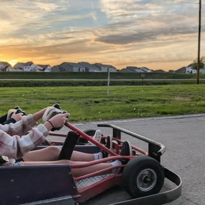 Two people drive a go-kart on a paved track at sunset at Boschertown Go Karts in St. Charles, with houses and a green field visible under a partly cloudy sky.