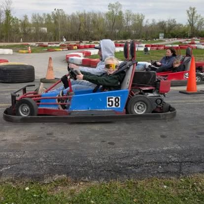 Two people ride a blue go-kart numbered 58 at Boschertown Go Karts in St. Charles, both wearing sunglasses. Another go-kart with two riders appears in the background, along with orange cones and trees lining the outdoor track.