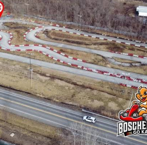 Aerial view of Boschertown Go Karts track in St. Charles, with winding red and white barriers bordered by brown grass and trees. A car drives on a road in the foreground. Boschetti Go Karting logo with cartoon mascot is in the corner.