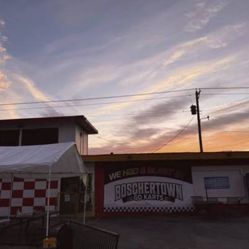 A sunset sky with wispy clouds above a St. Charles building displaying a sign that reads “Boschertown Go Karts” and “We had a blast!” A red-and-white checkered awning and tent are visible in the foreground.