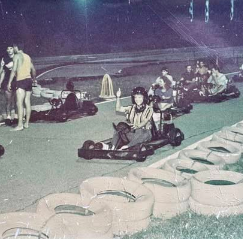 A person in a helmet and striped shirt gives a thumbs-up while sitting in a go-kart on the Boschertown Go Karts track at night in St. Charles. Several other people in go-karts and a few standing figures are visible in the background.
