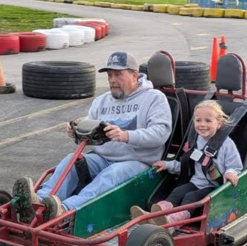 An older man and a young girl ride together in a go-kart on an outdoor track at Boschertown Go Karts in St. Charles. The man steers while the smiling girl sits beside him, with tires and traffic cones lining the track’s edge.
