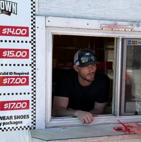 A man in a black shirt and cap leans out of a Boschertown Go Karts food stand window in St. Charles. A price board on the left lists ticket prices from $14 to $17. The man is holding a red pen and looking outside.