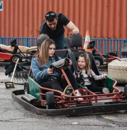 A man stands behind a go-kart while a girl drives and a younger child sits in the passenger seat at Boschertown Go Karts in St. Charles. Outdoors, other go-karts and traffic cones are visible in the background.