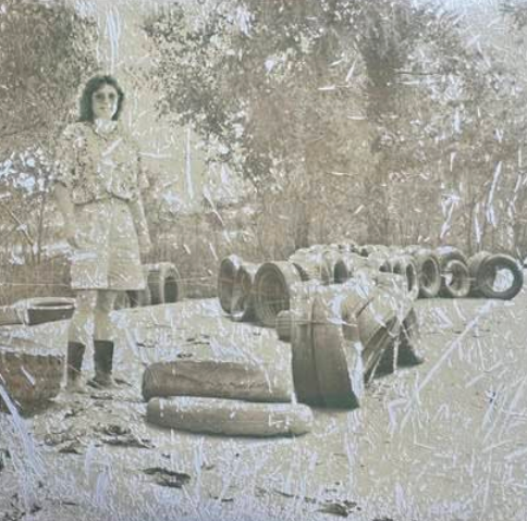 A woman stands outdoors beside scattered tires on the ground at Boschertown Go Karts. The sepia-toned, heavily scratched image has a worn, vintage look, with trees visible in the background of this St. Charles scene.