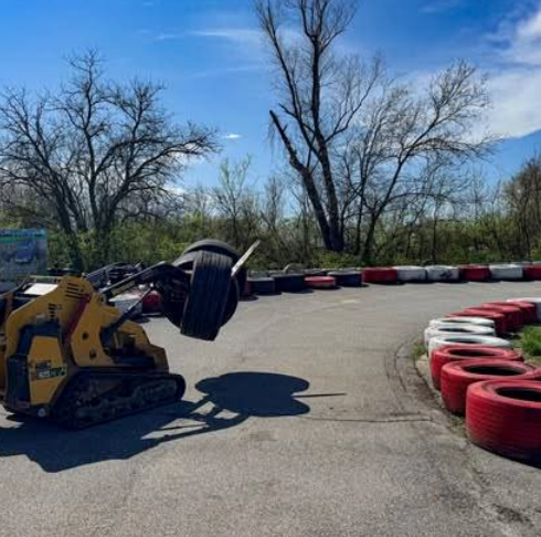A small yellow loader lifts several stacked tires on the outdoor Boschertown Go Karts track in St. Charles, lined with red, white, and black barriers, with trees and a blue sky in the background.
