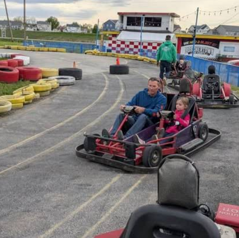 A man and a young girl ride together in a red go-kart at Boschertown Go Karts in St. Charles. Other go-karts and people are visible in the background, with colorful tires lining the outdoor track.