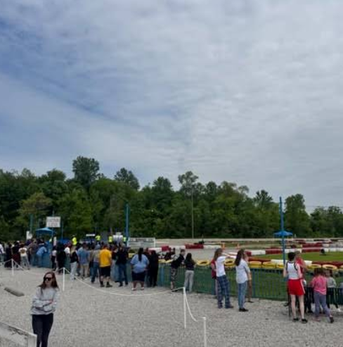 A group of people stands behind a white rope fence at Boschertown Go Karts in St. Charles, watching the go-kart track on a cloudy day. Trees line the background while a person in sunglasses and a gray outfit stands alone in the foreground.