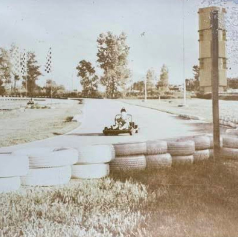 A person driving a go-kart on the outdoor Boschertown Go Karts track in St. Charles, surrounded by tire barriers and grass, with trees and checkered flags in the background. The image has a vintage, sepia tone.