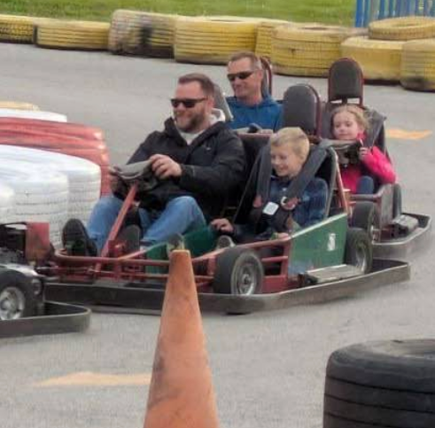 Two adults and two children enjoy an exciting race at Boschertown Go Karts in St. Charles, each adult driving with a child as a passenger. Tires and safety barriers surround the outdoor track for added safety.