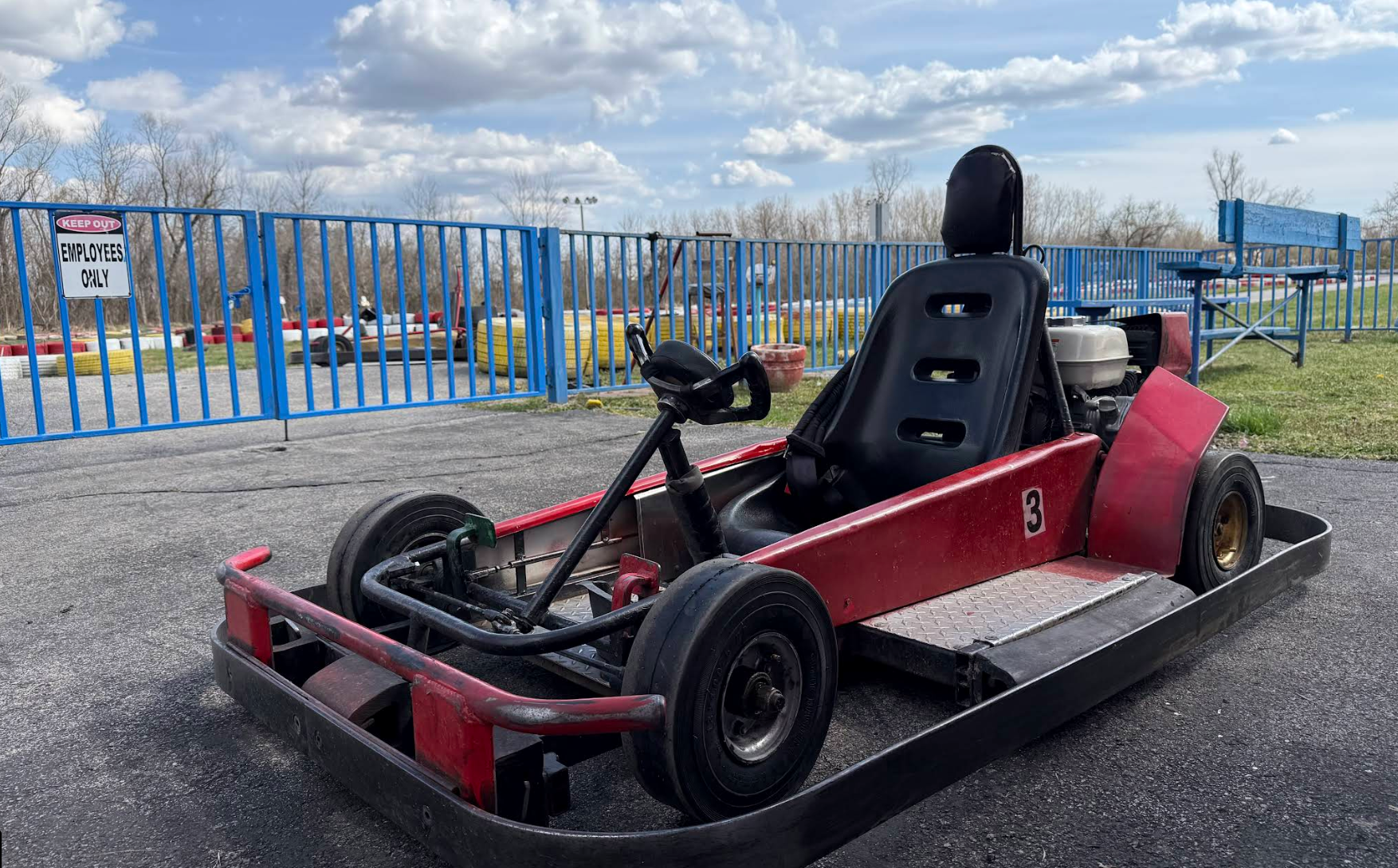 A red go-kart with the number 3 is parked on asphalt near a blue metal fence at Boschertown Go Karts in St. Charles. A sign on the fence reads EMPLOYEES ONLY, with trees and a racetrack visible under a partly cloudy sky.