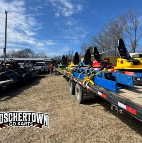 Several go-karts, secured with straps, sit on a flatbed trailer in an outdoor lot under a blue St. Charles sky. The Boschertown Go Karts logo is visible in the bottom left corner. Trees and vehicles are in the background.