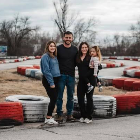 Four people, including a man, two women, and a young girl, smile together by the Boschertown Go Karts track in St. Charles, surrounded by red and white tires, with leafless trees and cloudy skies in the background.
