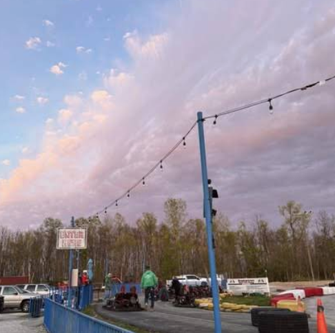 A go-kart track at sunset in St. Charles, with people near Boschertown Go Karts, string lights overhead, trees in the background, and an ENTRY sign on the left. The sky is filled with pastel clouds.