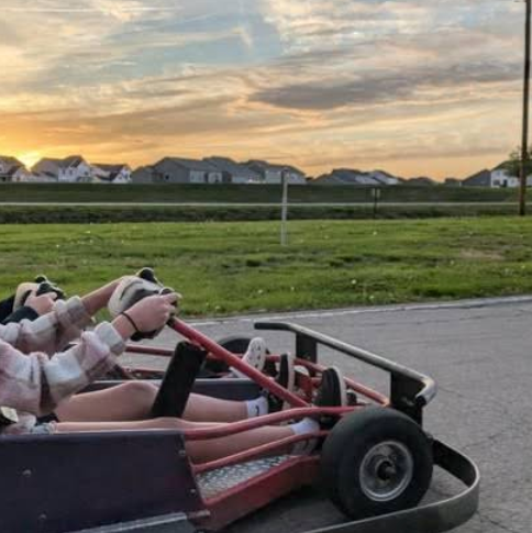 Two people drive a go-kart on the paved outdoor track at Boschertown Go Karts in St. Charles at sunset, with grassy fields and houses visible in the background. The sky is partly cloudy, and both drivers are wearing gloves.
