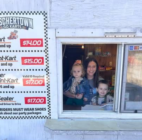 A woman and two young children stand at a wooden outdoor ticket window at Boschertown Go Karts in St. Charles. A price sign for go-kart rides is visible on the left, listing rates and rules. The window is part of a white building.