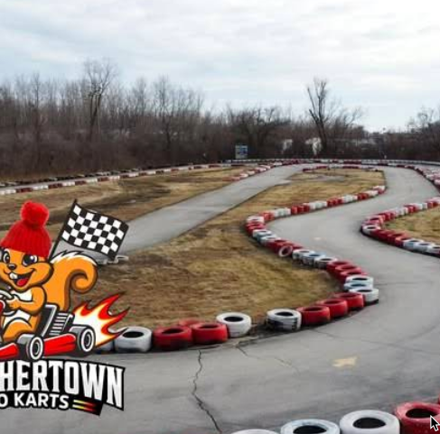 A winding outdoor go-kart track bordered by red and white tires weaves through a grassy area with bare trees in St. Charles. A cartoon squirrel holding a racing flag and the text “SQUIRRELTOWN GO KARTS” sits in the lower left corner.