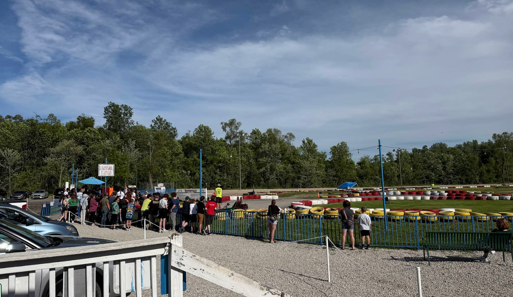A group of people waits in line near the Boschertown Go Karts track in St. Charles, surrounded by trees and colorful tire barriers under a partly cloudy sky. Some watch the go karts from behind a green fence.