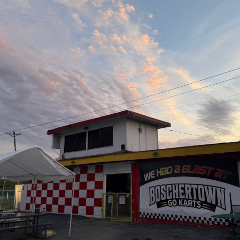 A go-kart building with red and white checkerboard walls sits under a sunset sky in St. Charles. The sign reads “We had a blast at Boschertown Go Karts.” A canopy and picnic table are in the foreground.