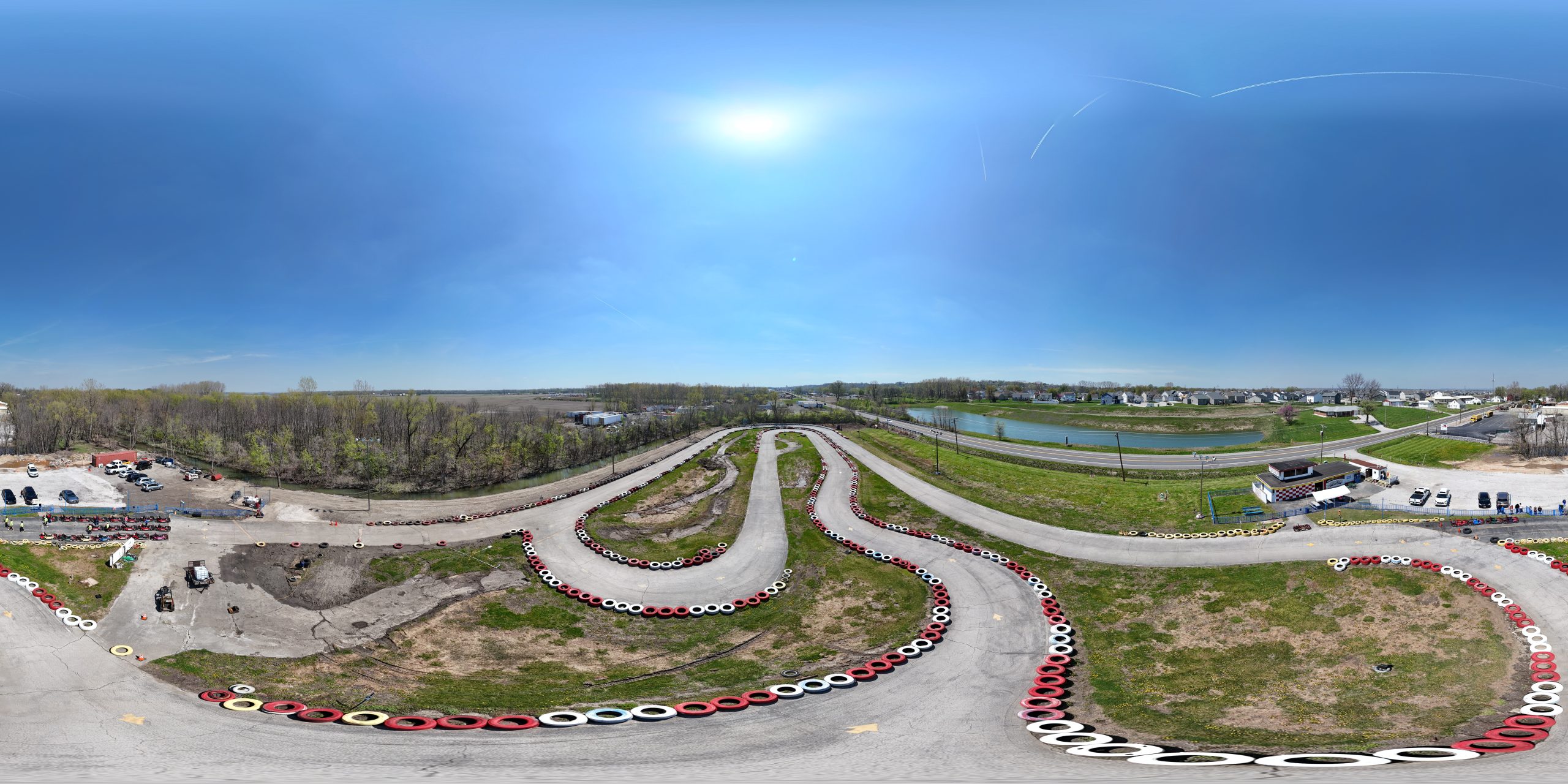 Aerial, 360-degree view of Boschertown Go Karts in St. Charles, featuring winding go-kart tracks bordered by red and white tires, grassy areas, trees, and a few buildings under a clear blue sky.