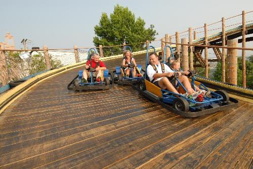 Four people ride Go Karts at Boschertown Go Karts in St. Charles on a wooden outdoor track, with trees and a fence in the background. The two karts are side by side as they round a curve, and the riders appear to be enjoying themselves.