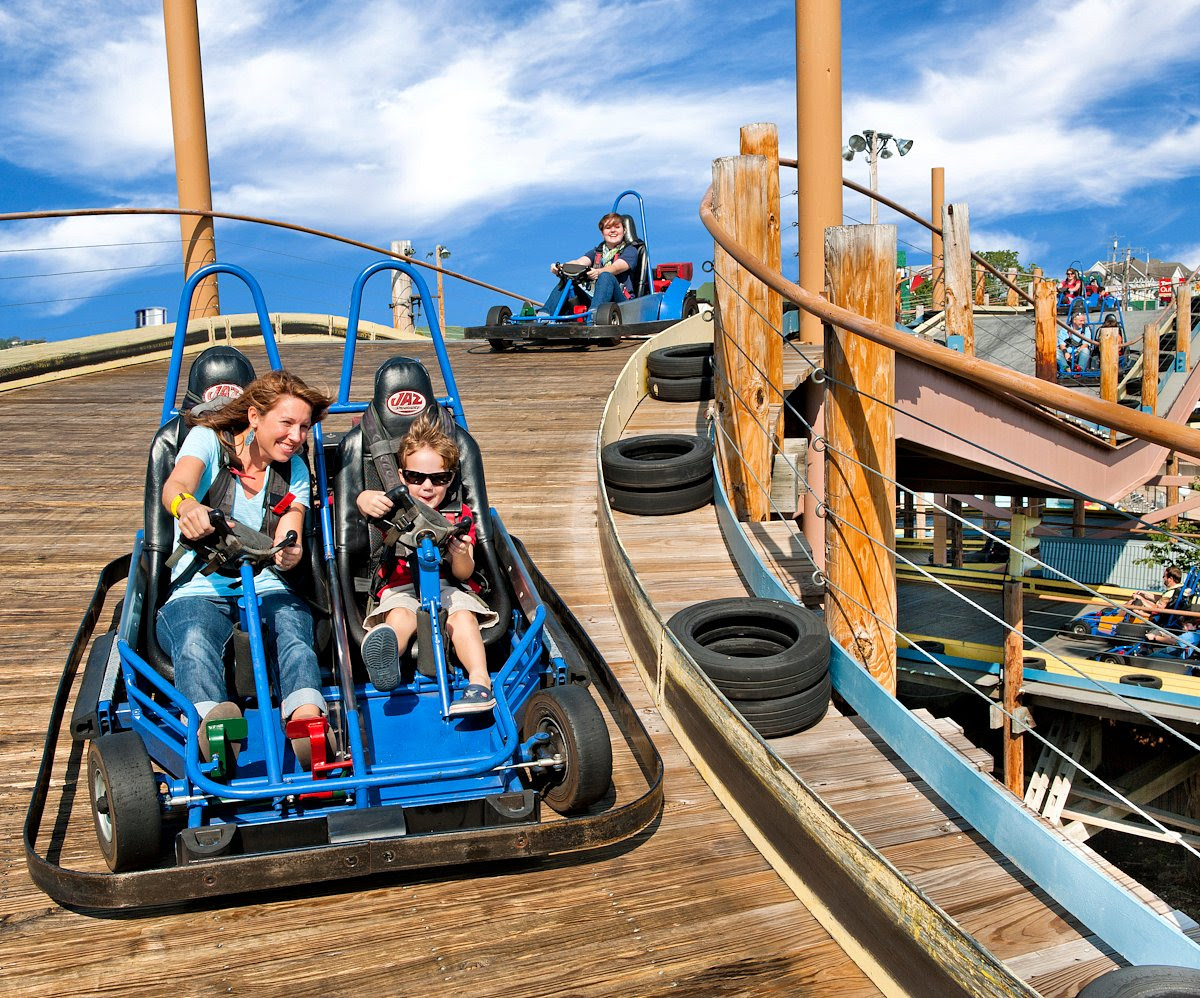 A woman and a child ride a blue go-kart down the wooden track at Boschertown Go Karts in St. Charles, both smiling as they steer. Other people follow behind under a bright blue sky with scattered clouds.