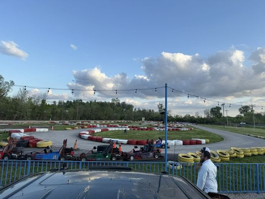 Go Karts lined up on the outdoor track at Boschertown Go Karts in St. Charles, with colorful tire barriers. Several people are seated in the go-karts, while others stand nearby amid trees and clouds in the background.